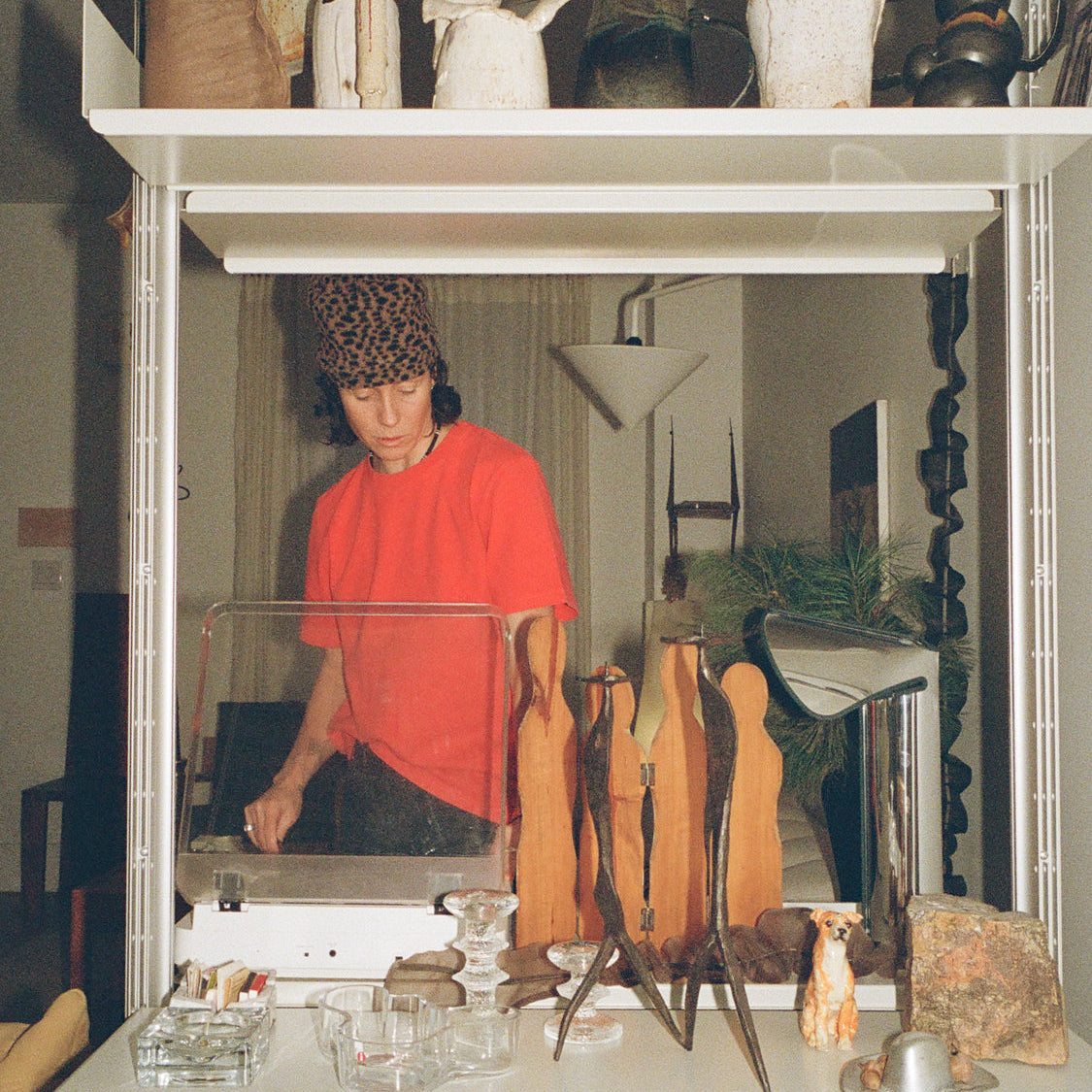 Person wearing a red shirt and patterned headscarf standing behind a display case filled with various objects.