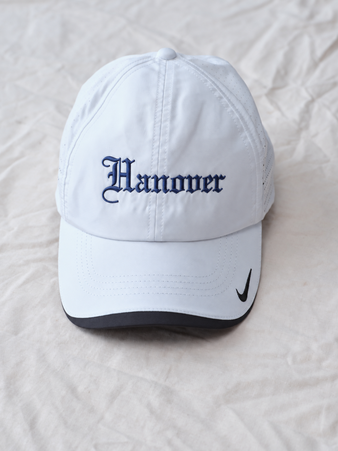 A white baseball cap with 'Hanover' embroidered in dark blue Gothic lettering on the front, set against a wrinkled off-white fabric background.