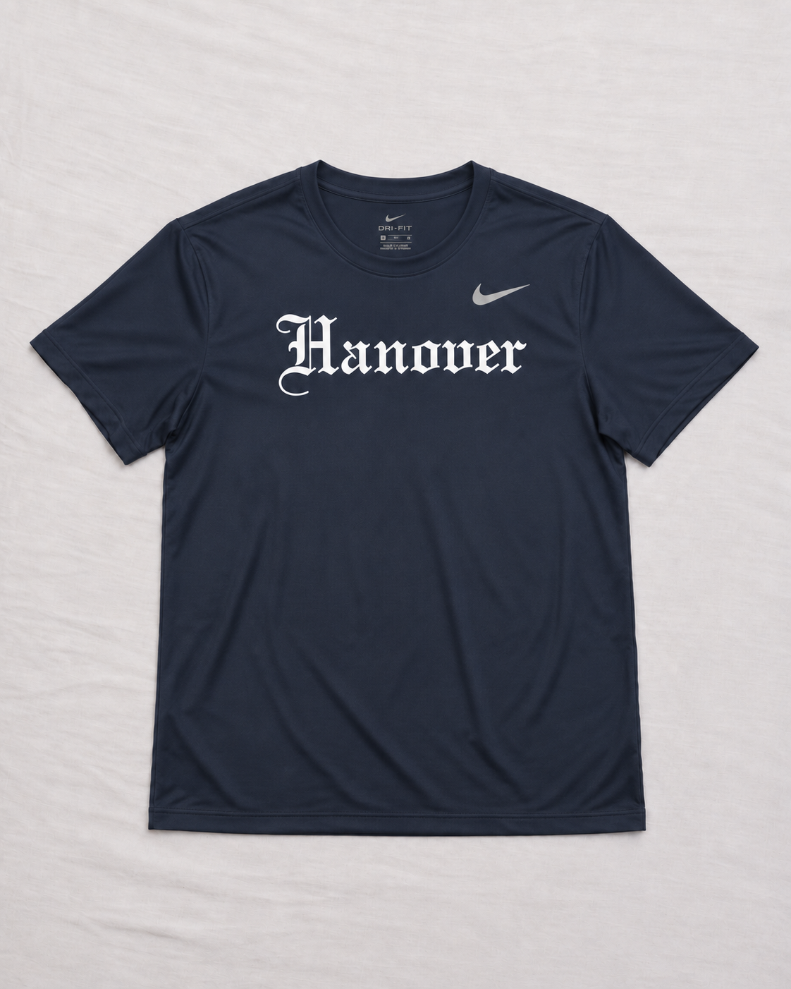 A navy blue Hanover athletic t-shirt with 'Hanover' printed in a white Old English font across the chest, laid flat on a light gray fabric background.