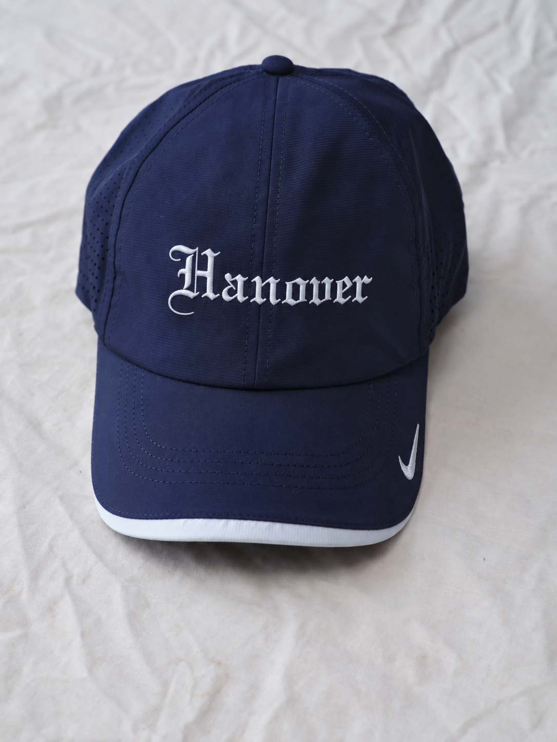 A navy blue baseball cap with 'Hanover' embroidered in white Gothic lettering on the front  resting on a wrinkled off-white fabric background.