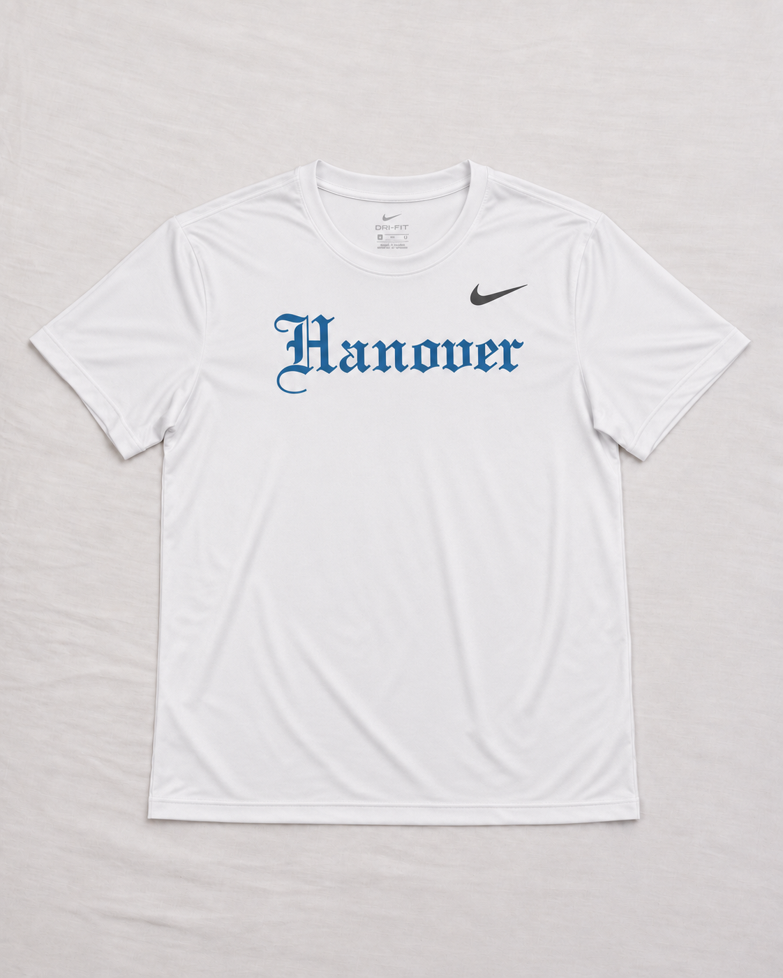 A white short-sleeve Hanover t-shirt with 'Hanover' printed in a blue Gothic font across the chest  laid flat on a light gray fabric background.