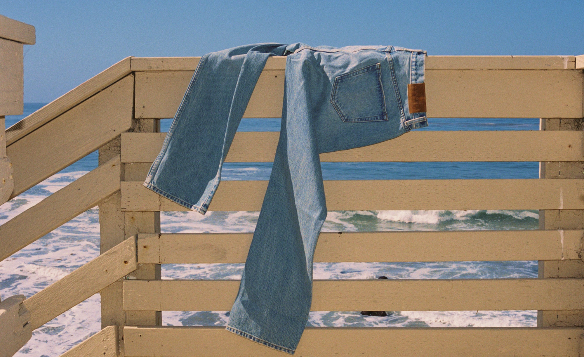 Hanover Drift Jeans jeans draped over a wooden railing with a beach and ocean view in the background
